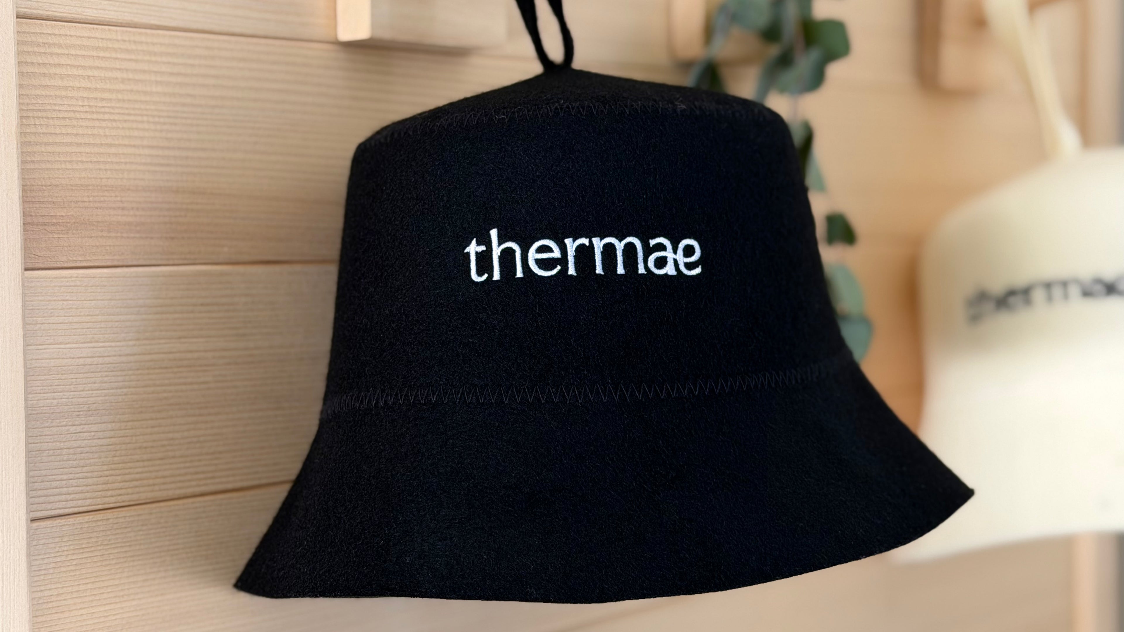 Black merino wool sauna hat with 'thermae' branding on a wooden surface. No synthetic materials.